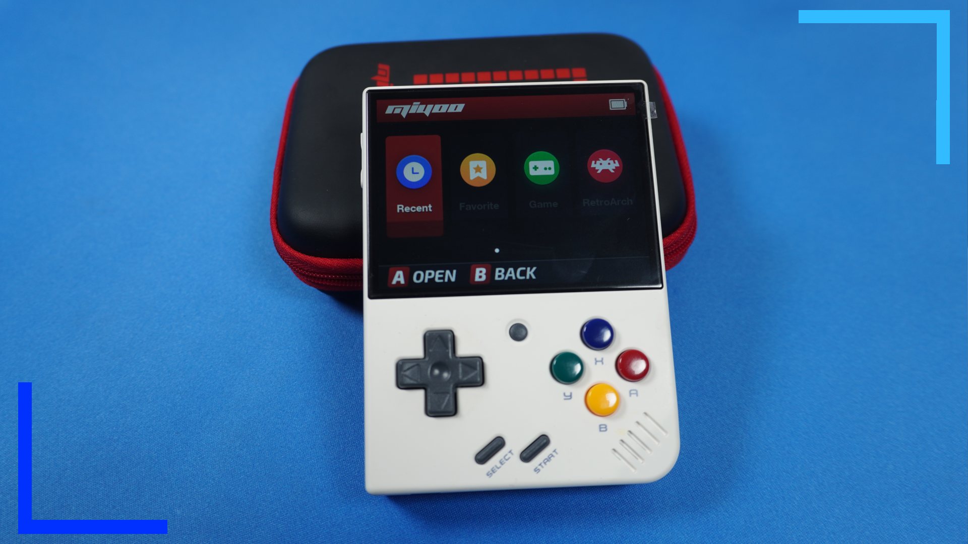 The Miyoo Mini rest up against the hard carry case. The Mini's screen shows four icons which point to games, RetroArch, favorites, and settings.